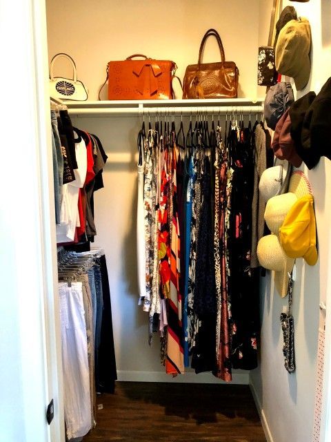 Small closet filled with hanging clothes, purses, and hats. Brown wood floor and white shelves.