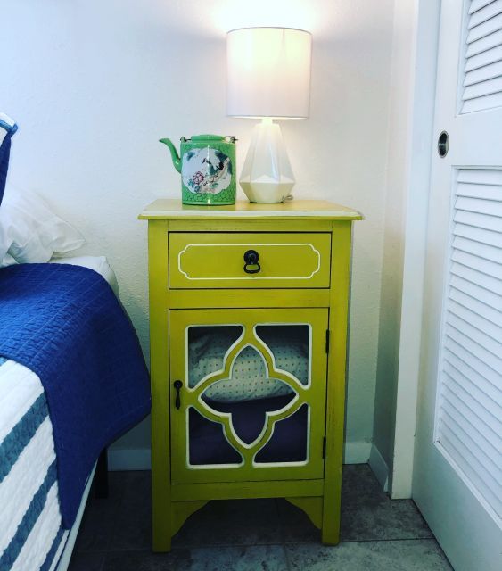 Lime green nightstand with lamp, teapot, and patterned door. Next to a bed.
