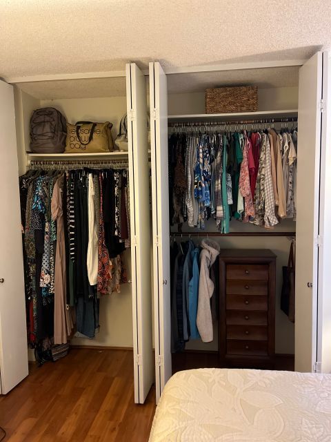 Closet with hanging clothes and shelves. White doors are open. Wooden floor, brown and white clothing.