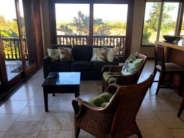 Living room with a dark sofa, wicker chairs with green cushions, and a view through large windows.