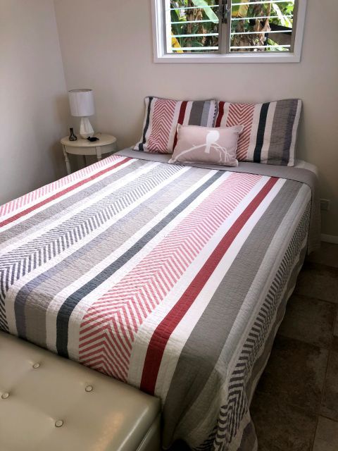 Bedroom with a bed covered in a striped red, gray, and white quilt, small bench at foot, a side table and window above.