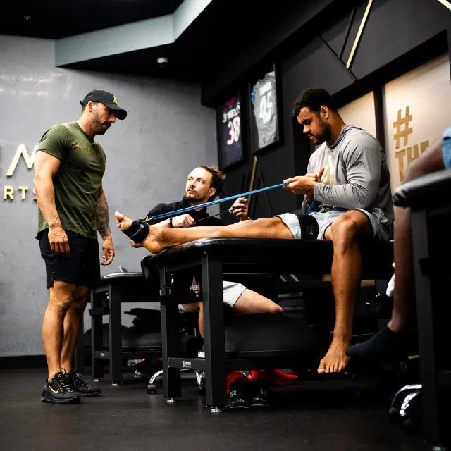 Three men in a gym setting. One man stretches a leg on a table using a resistance band, while another watches and assists.