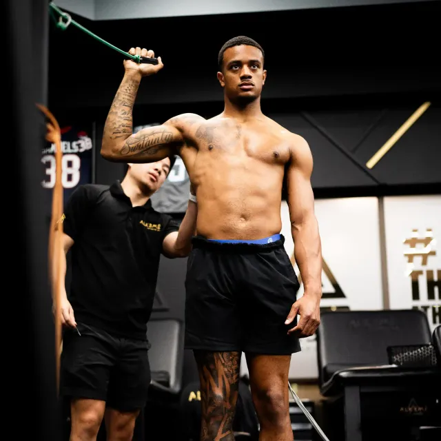 Man exercising shoulder with trainer in a gym setting, using a resistance band.