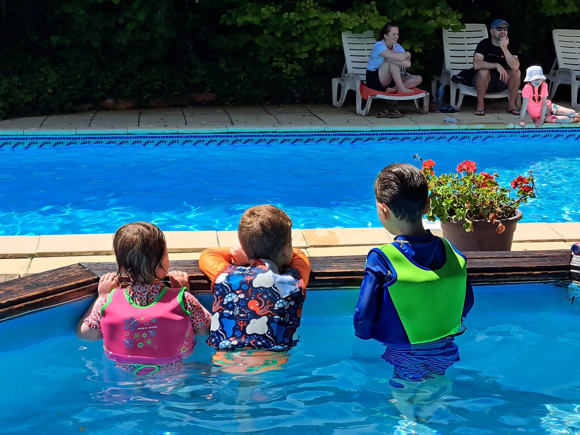 three children stood in a small childrens pool looking away over the edge at a bigger swimming pool