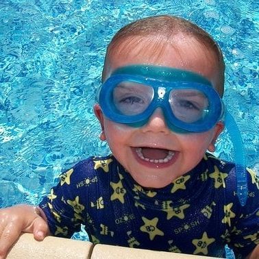 a child in a pool wearing goggles and grinning at the side of a pool