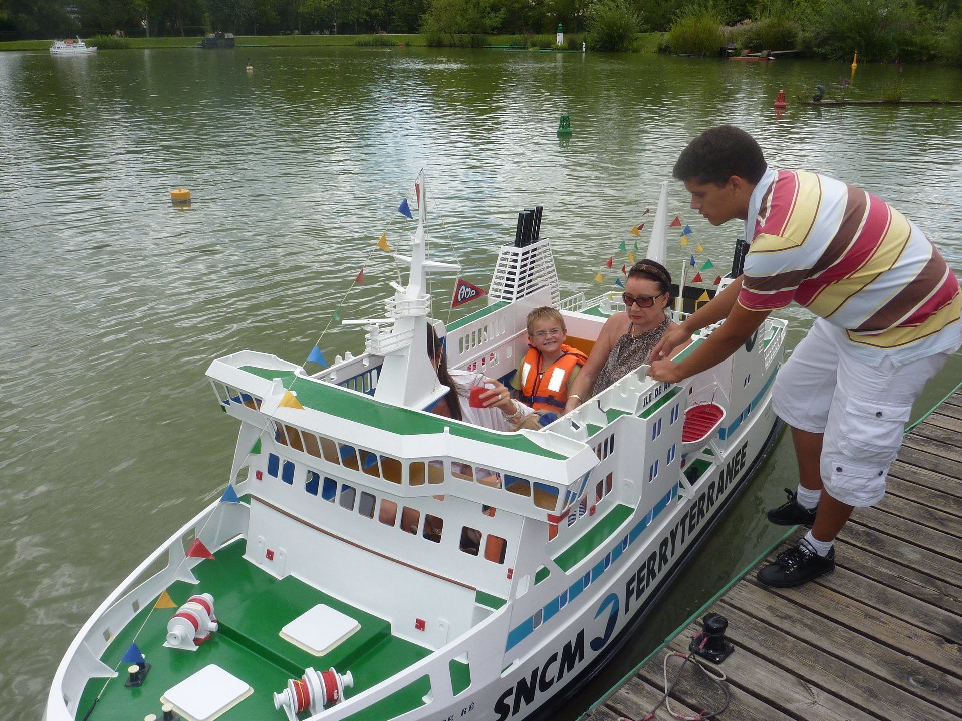 an adult and child sat in an electric boat shaped and decorated like a ferry. The child is wearng a life jacket