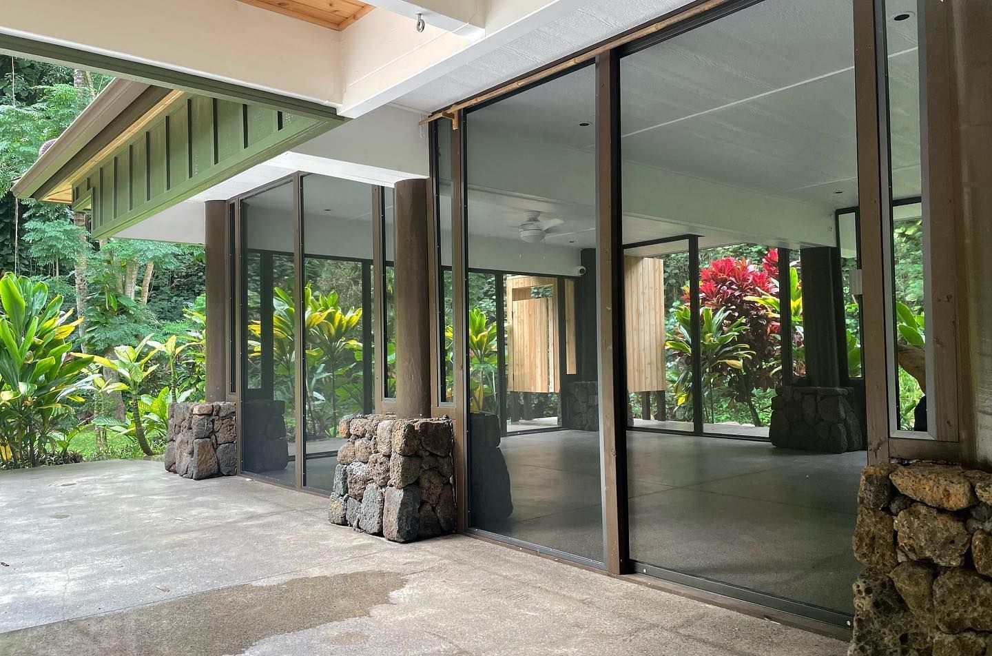Exterior of a building with glass walls, concrete pillars, and a shaded roof. Lush green plants surround it.