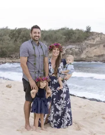 Family of four on a beach. Woman in floral dress holds a baby, daughter in a blue dress, man in gray shirt.