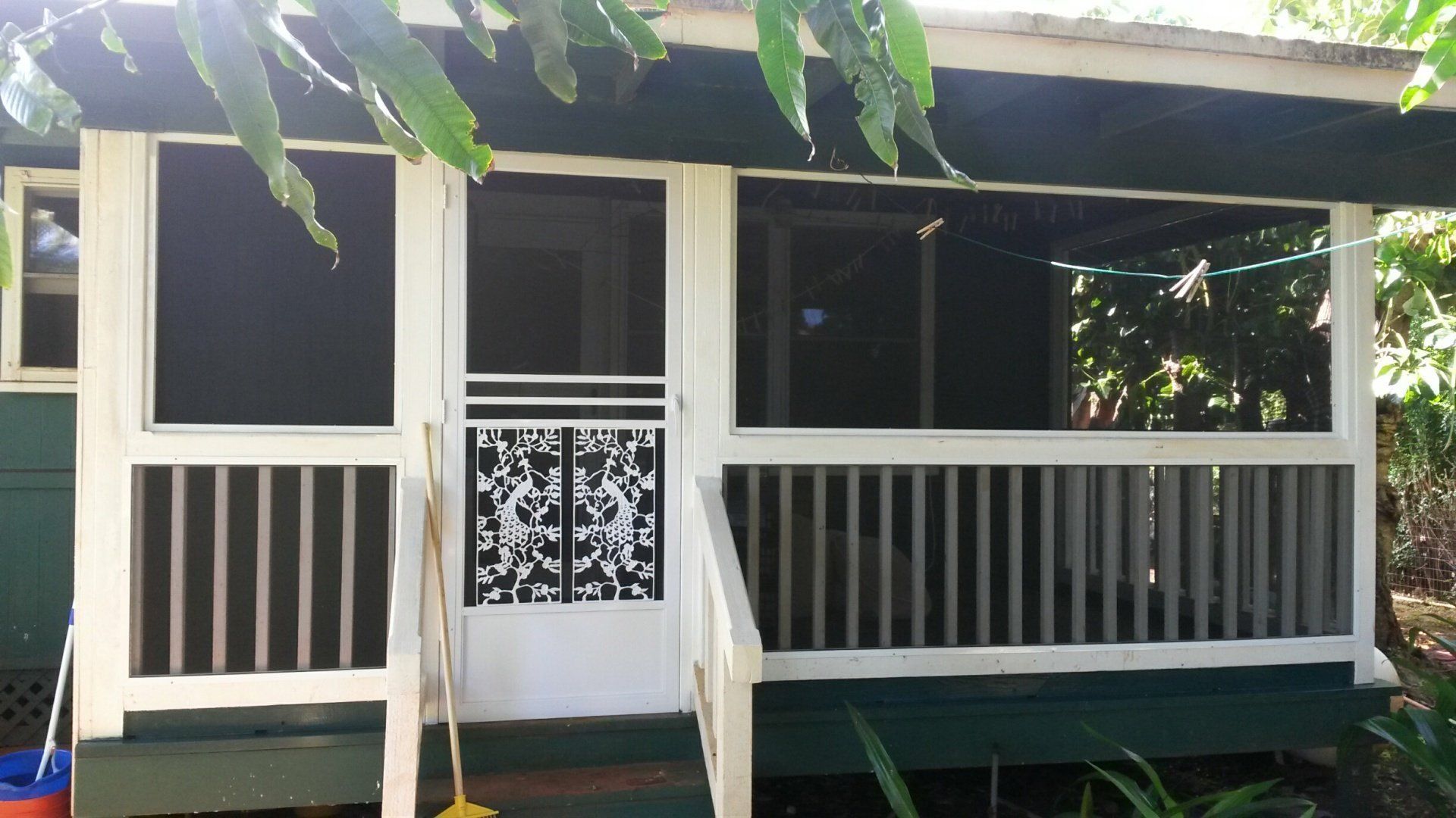 A screened-in porch with a white railing and door. Green house with plants.