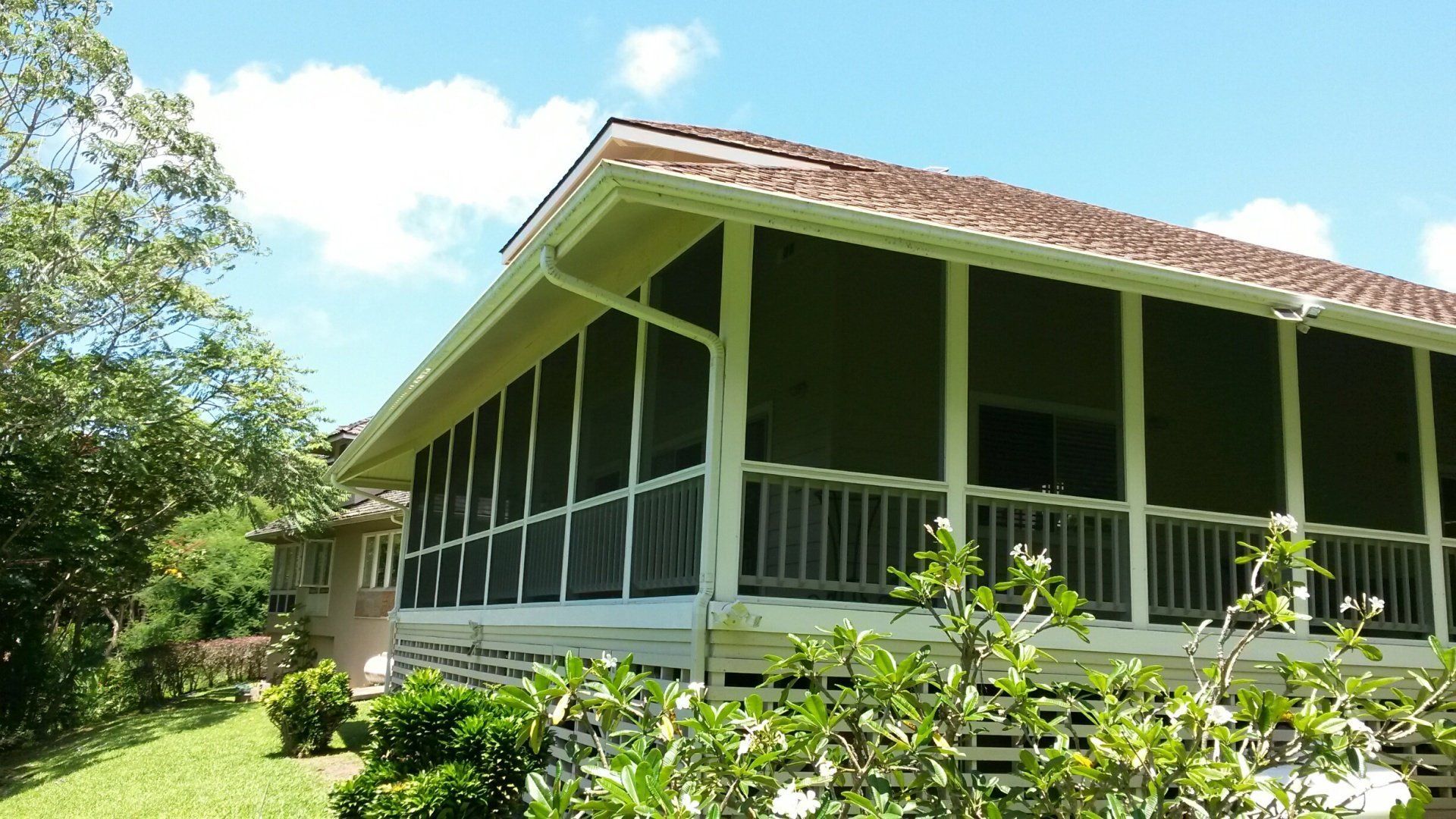 Screened porch with white railings and gutters under a brown roof, surrounded by green foliage, blue sky.