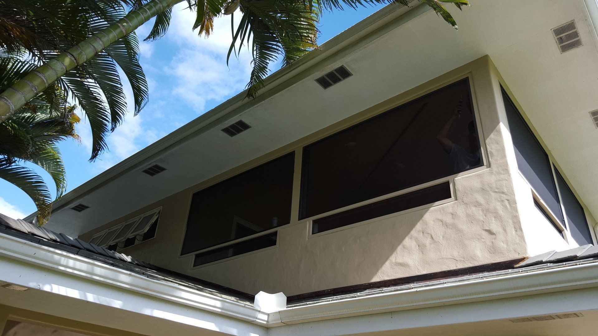Exterior view of a house with large screened windows, beige walls, and a white roof against a blue sky.