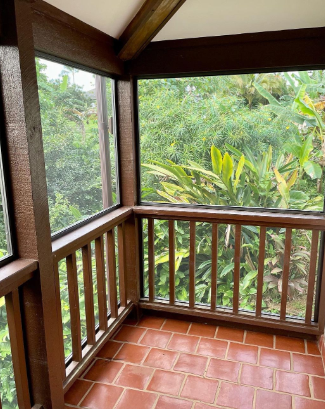 Screened porch with brown railing and floor tiles, overlooking lush green foliage.