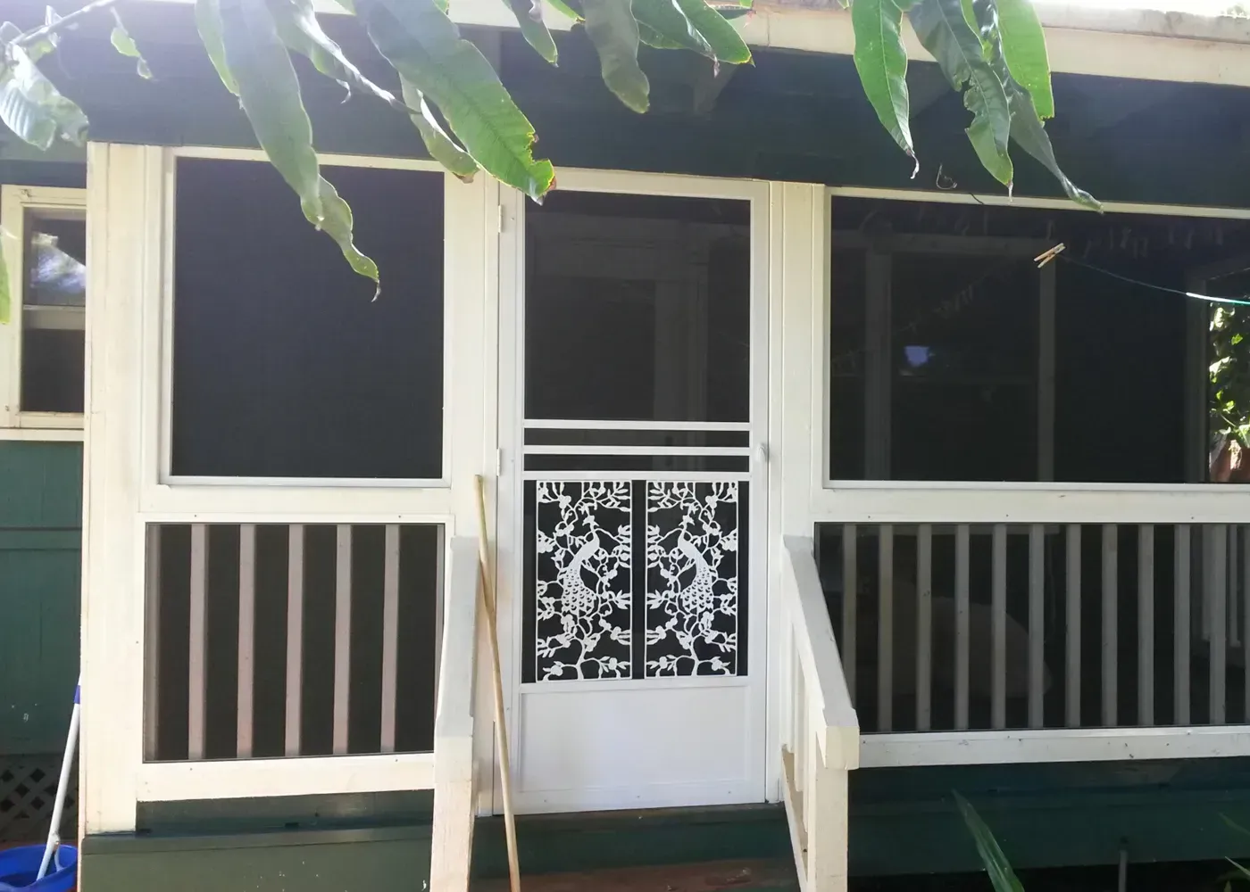 White screen door and porch with railings. Green siding, leafy branches overhead.