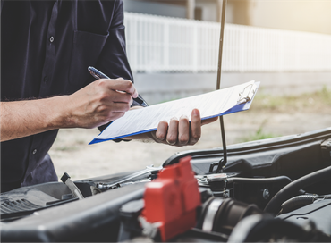 A man is writing on a clipboard while looking under the hood of a car.