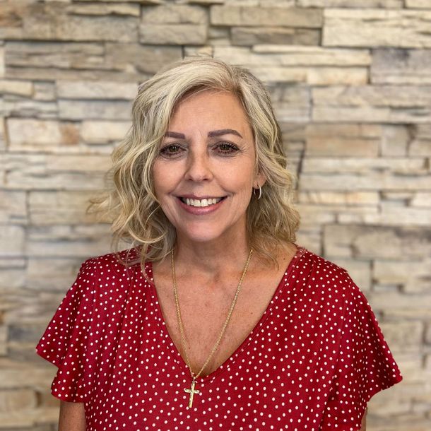 Woman with blonde wavy hair, wearing a red polka dot top, smiling, necklace with cross. Against a brick wall.