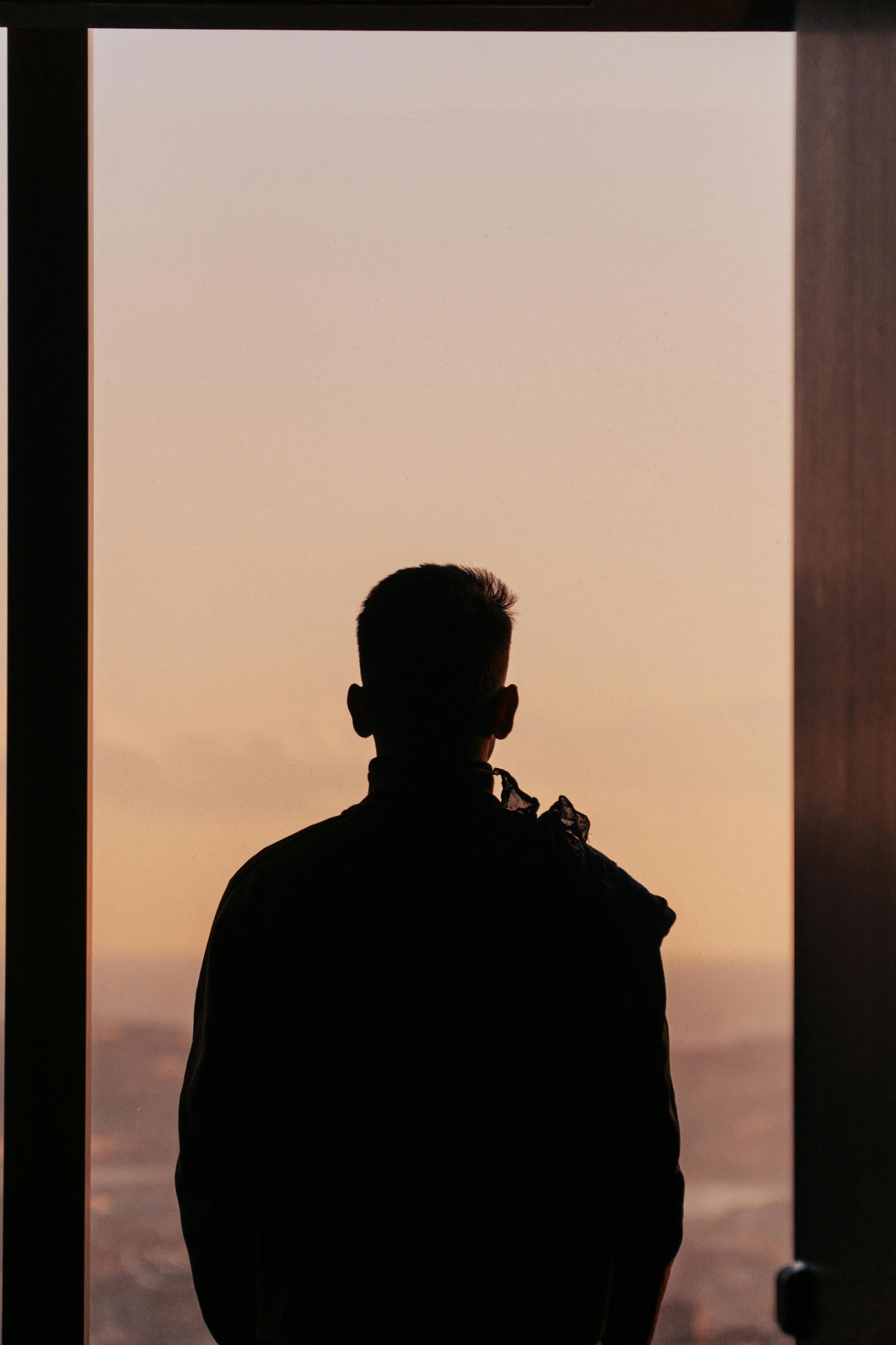 Silhouette of a person looking out a window at a sunset with orange and pink hues.