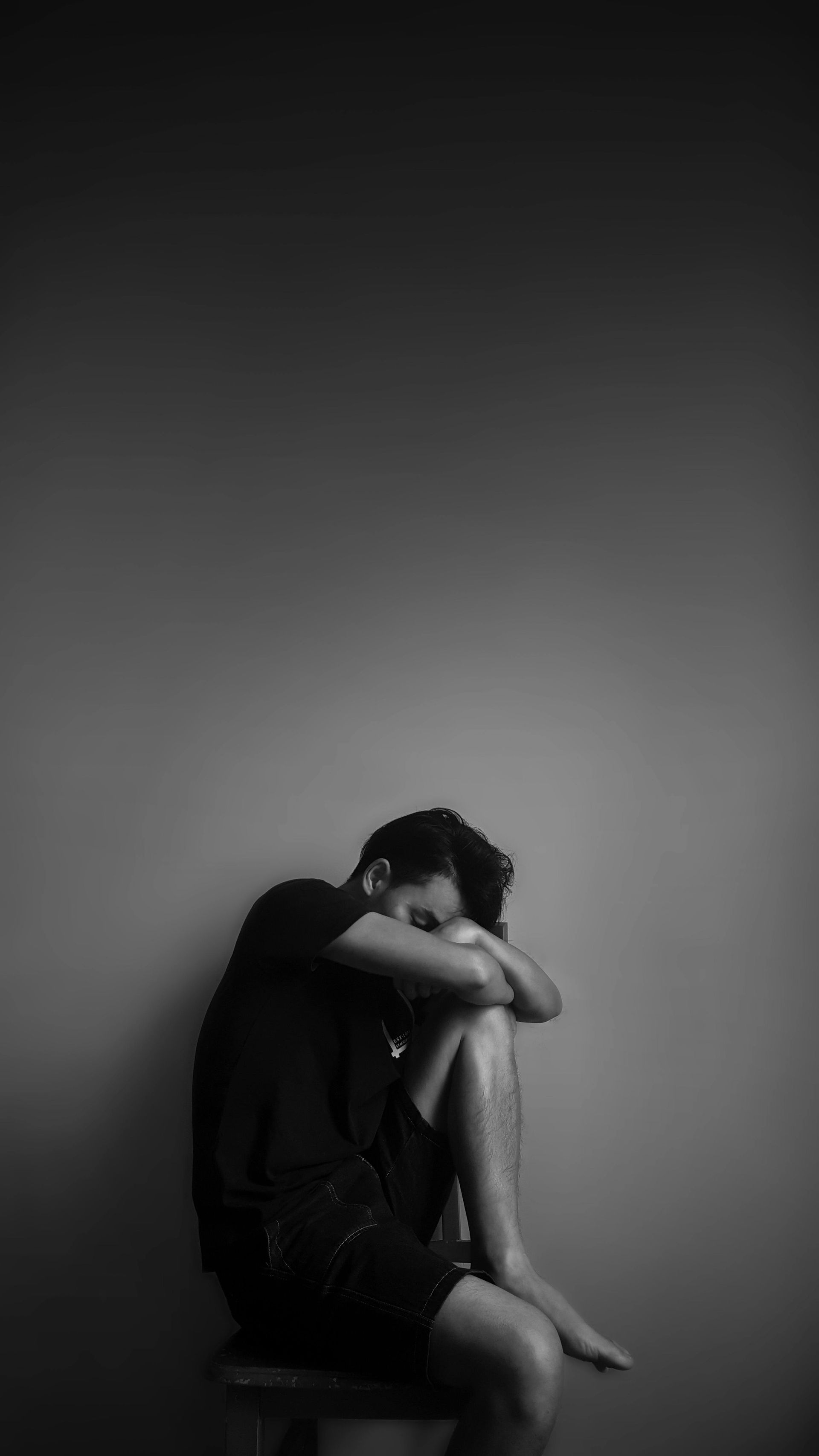 Man sits hunched over, head down, knees drawn up, against a gray wall. Black and white.