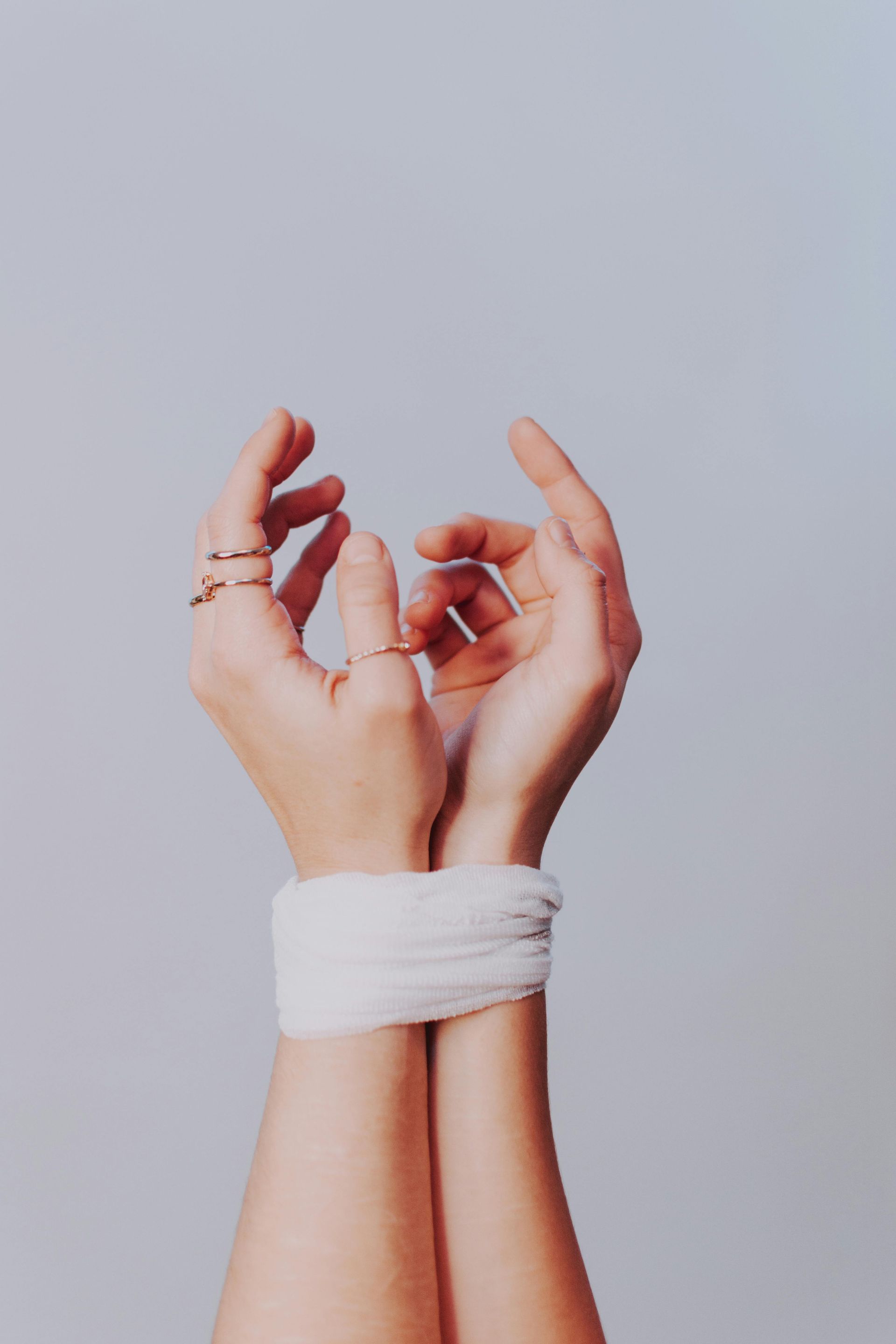 Two hands, wrists bound with white cloth, reach upward, fingers adorned with rings, pale gray background.
