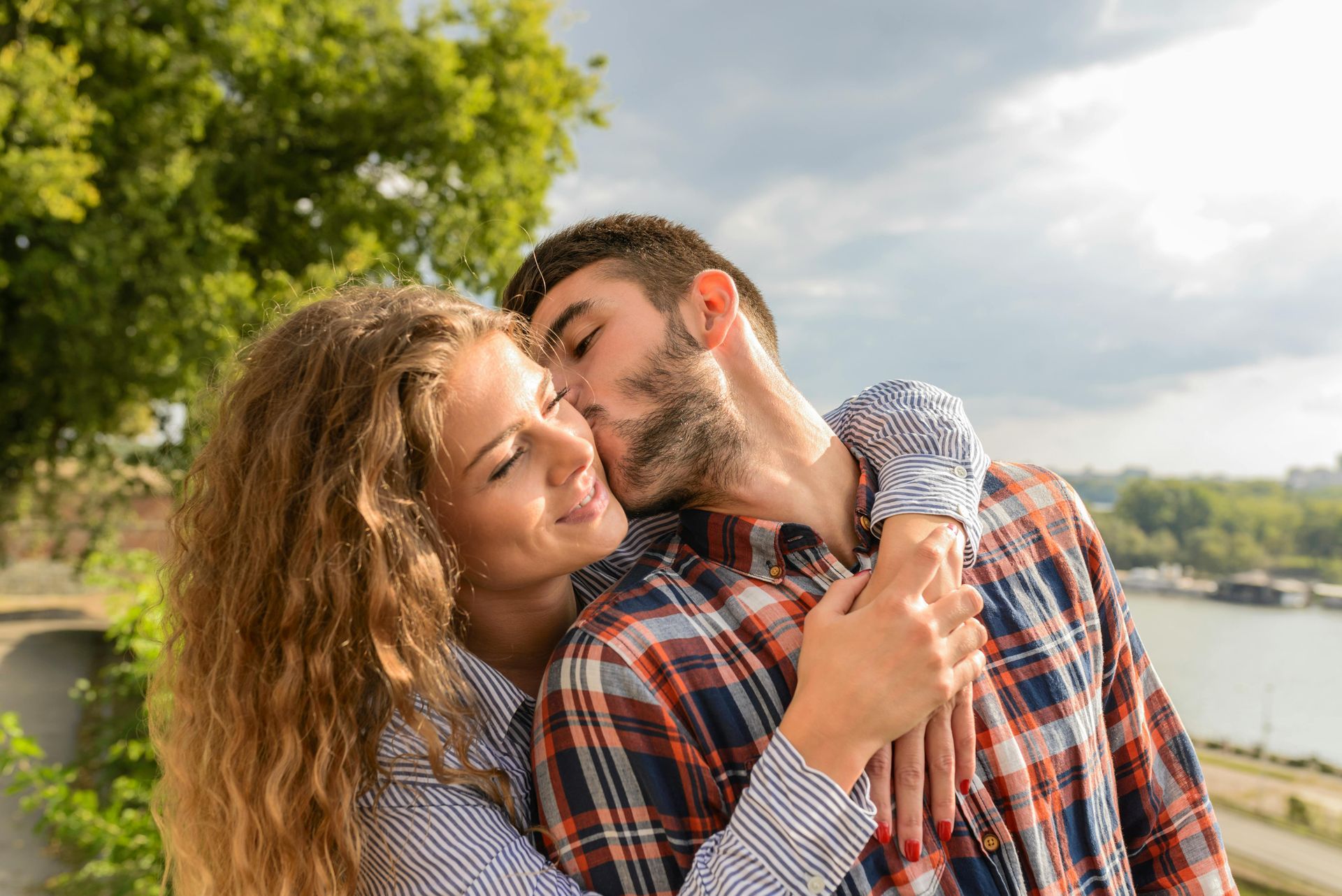 Man kisses woman's cheek outdoors; they embrace with a river and trees in the background.