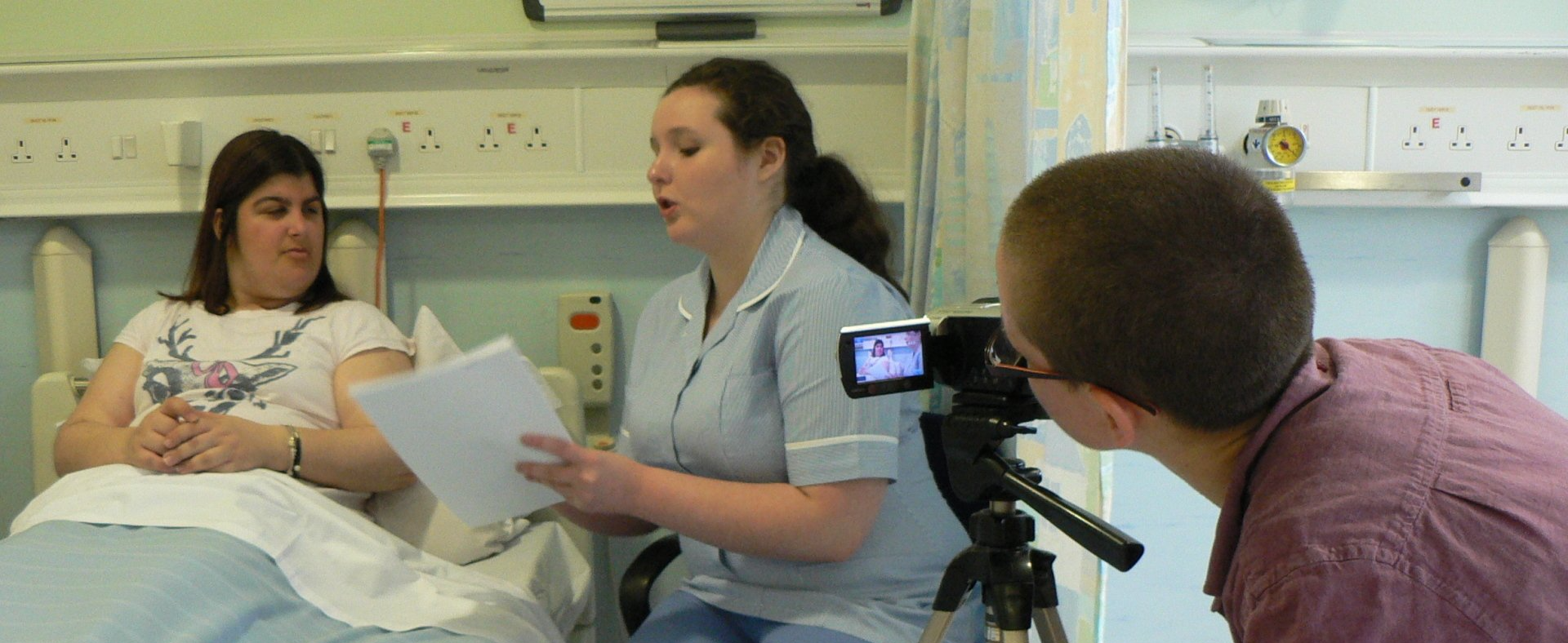 A woman in a hospital bed with a nurse sitting next to the bed, reading from a document. In the foreground is a person behind a camera, filming the scene