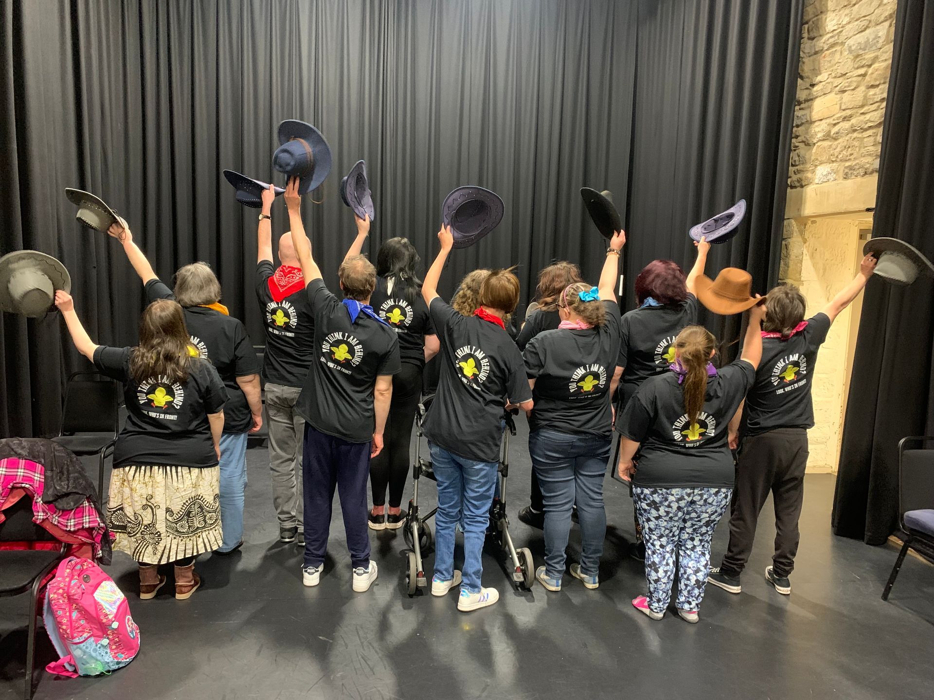 Group of performers stand on stage with their back to the audience wearing black t shirts and cowboy hats