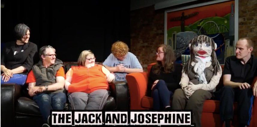 7 people sitting on or near 2 settees. One person is in a costume with a large mask on their head. The photo is called The Jack and Josephine