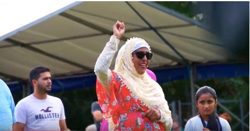 A woman wearing a hijab and sunglasses, dancing with one arm held high. Other people dancing in the background.