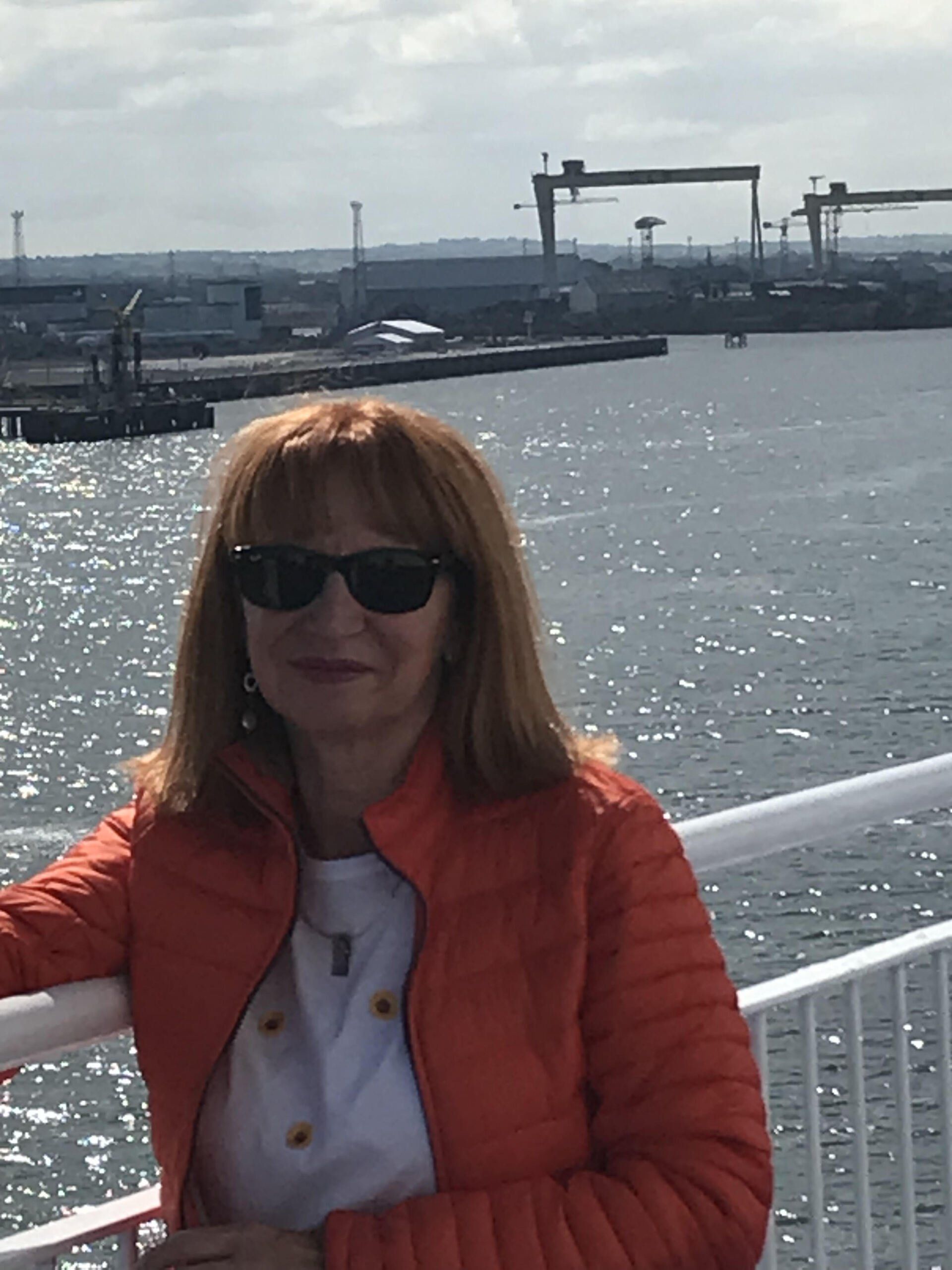 Una, with shoulder length hair and wearing sunglasses. She is standing next to railings with water and a ship yard in the background.