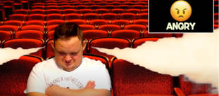 A young man with learning disabilities sitting on a seat in a theatre, with his arms crossed and looking sad and angry. There is an angry emoji in the top right corner of the image
