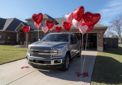 Silver pickup truck parked in a driveway with red heart balloons tied to it.