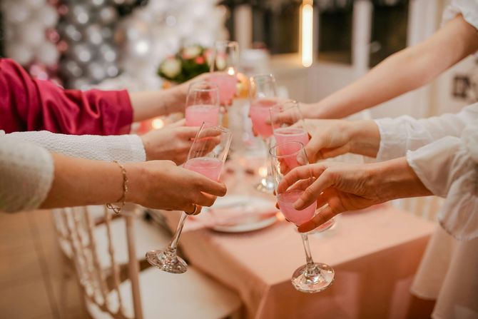 Hands clinking champagne flutes filled with pink liquid, celebrating around a table.