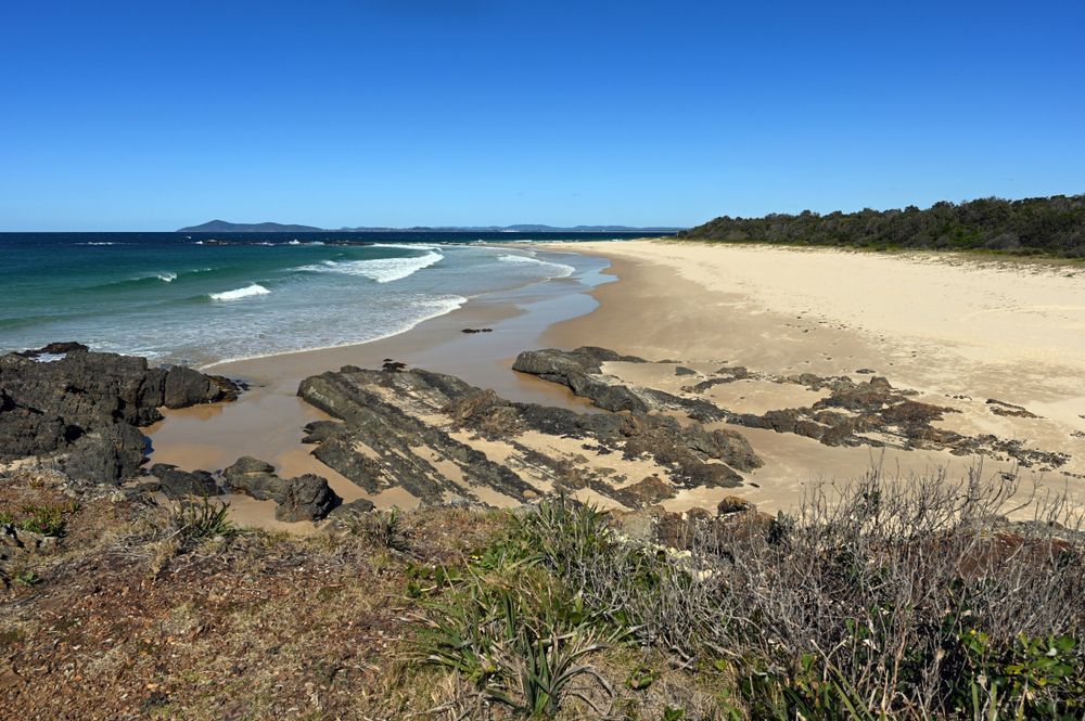 Beach Scene With Sand, Waves, and Rocky Shoreline Under a Blue Sky — CP Roofing in Hallidays Point, NSW
