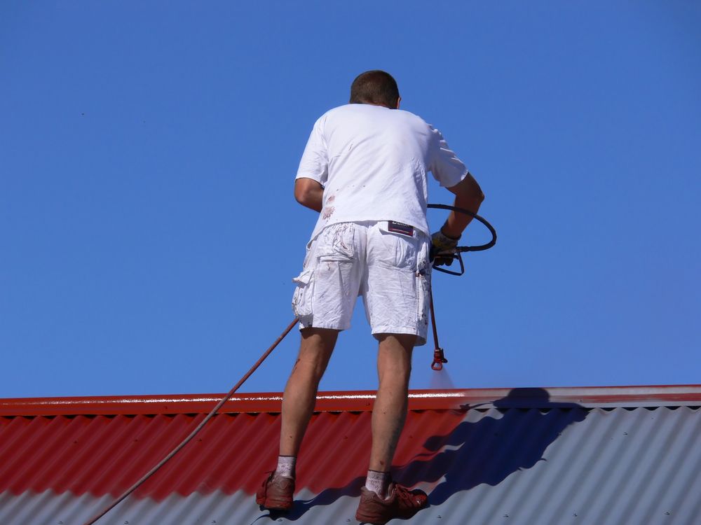Man in Shorts Spray-painting a Red Roof Against a Clear Blue Sky — CP Roofing in Old Bar, NSW