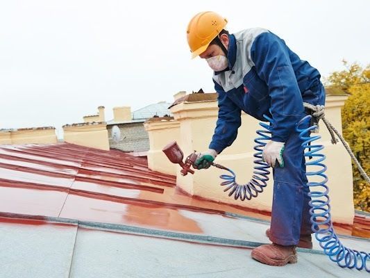 A Man Wearing A Hard Hat And A Mask Is Spraying Paint On A Roof — CP Roofing in Taree, NSW