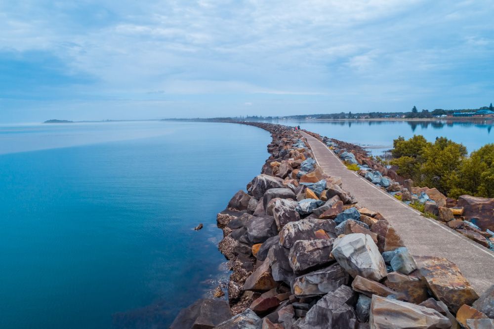 A Concrete Walkway Surrounded By Rocks Leading To A Body Of Water — CP Roofing in Harrington, NSW