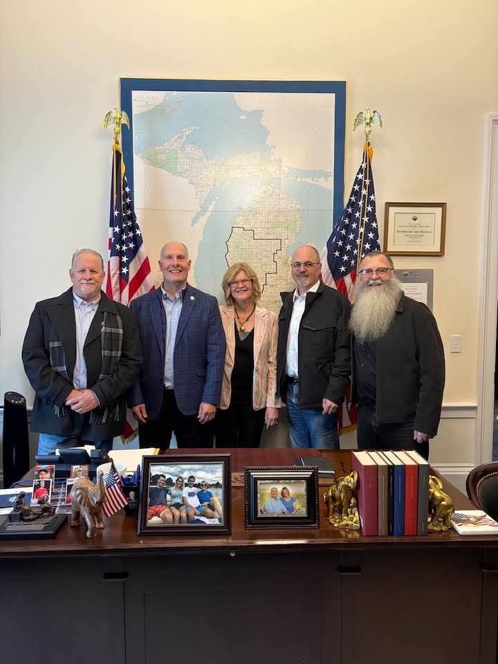 Five people posing together in an office, in front of a desk, American flags and a map of Michigan in the background.