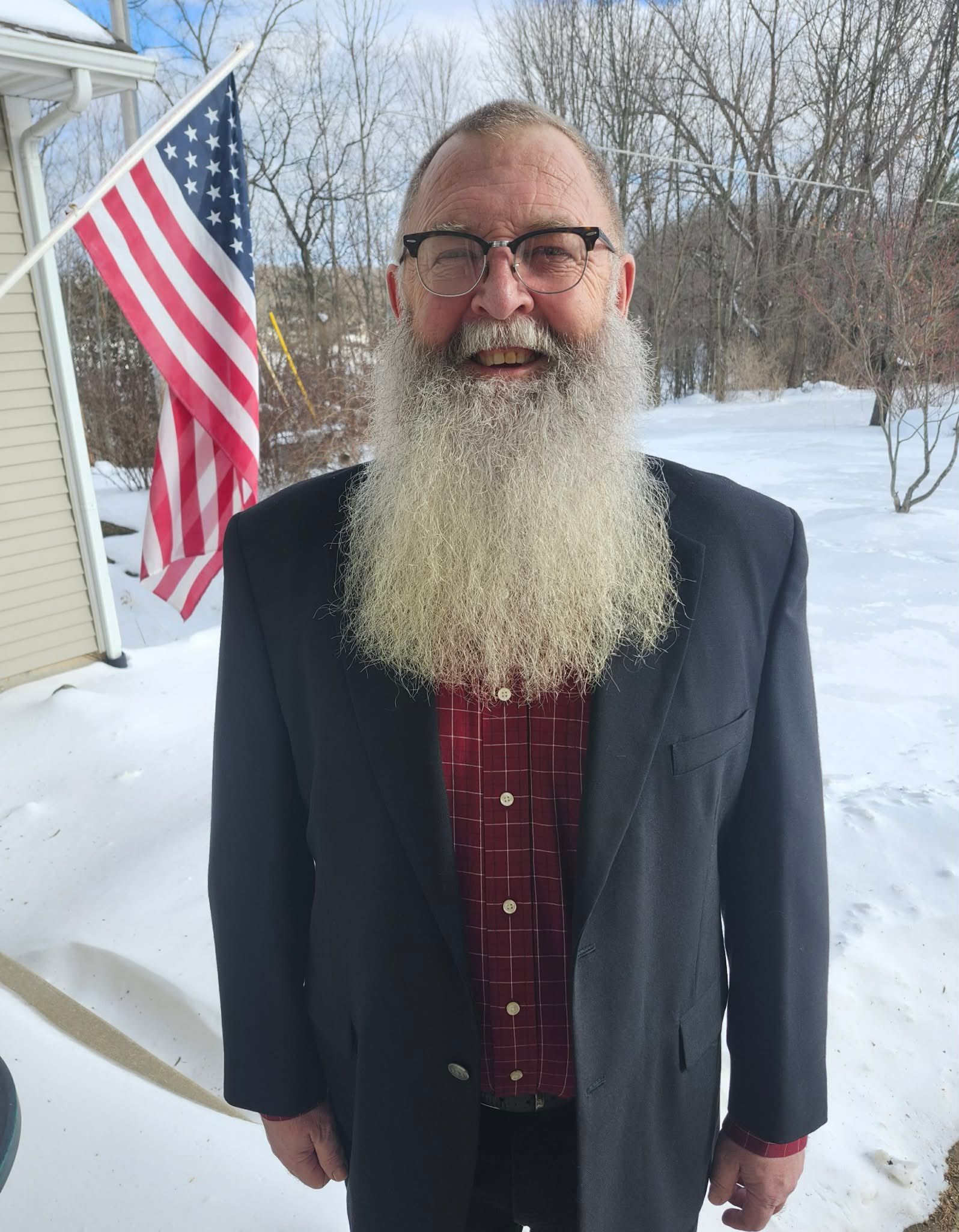 Man with long, white beard, wearing glasses, blazer, and red shirt, stands in front of a snow-covered yard with an American flag.