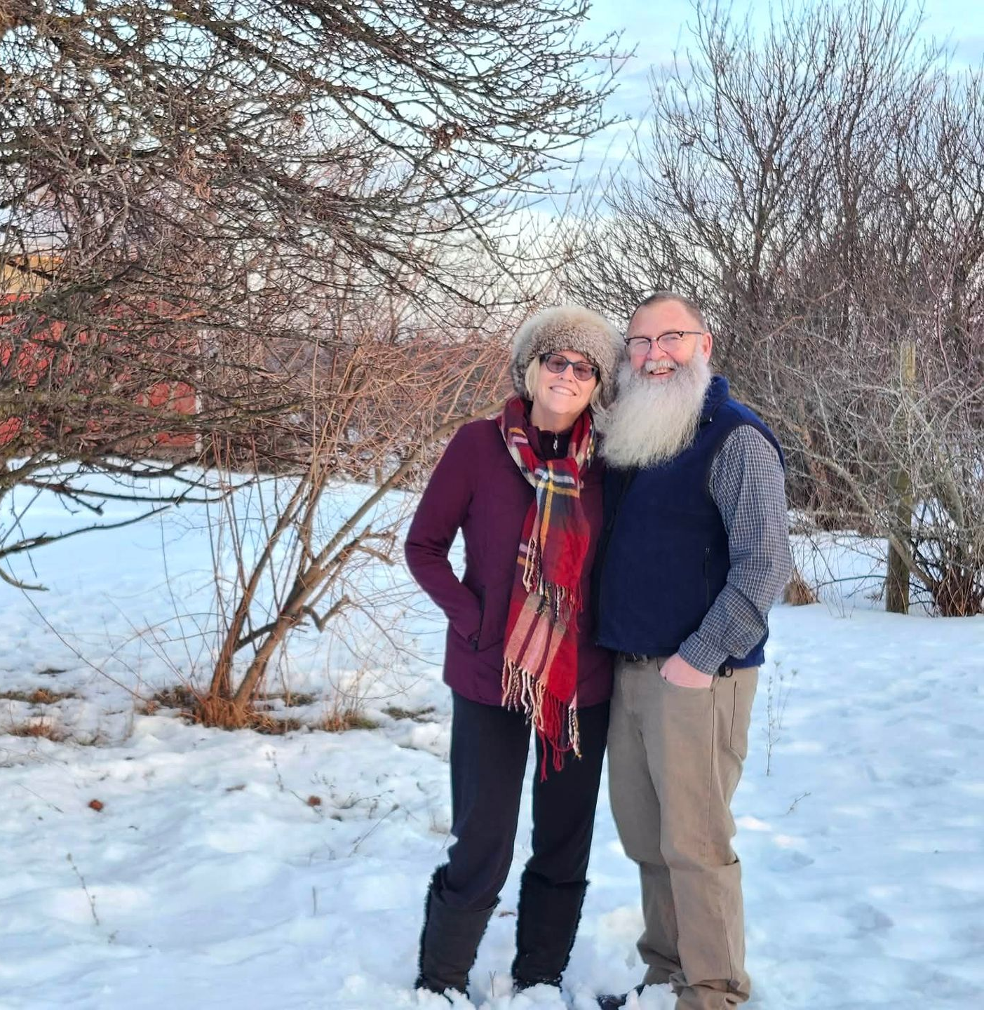 Couple smiles in snowy setting, man with long beard, woman in fur hat and scarf, trees and red building in background.