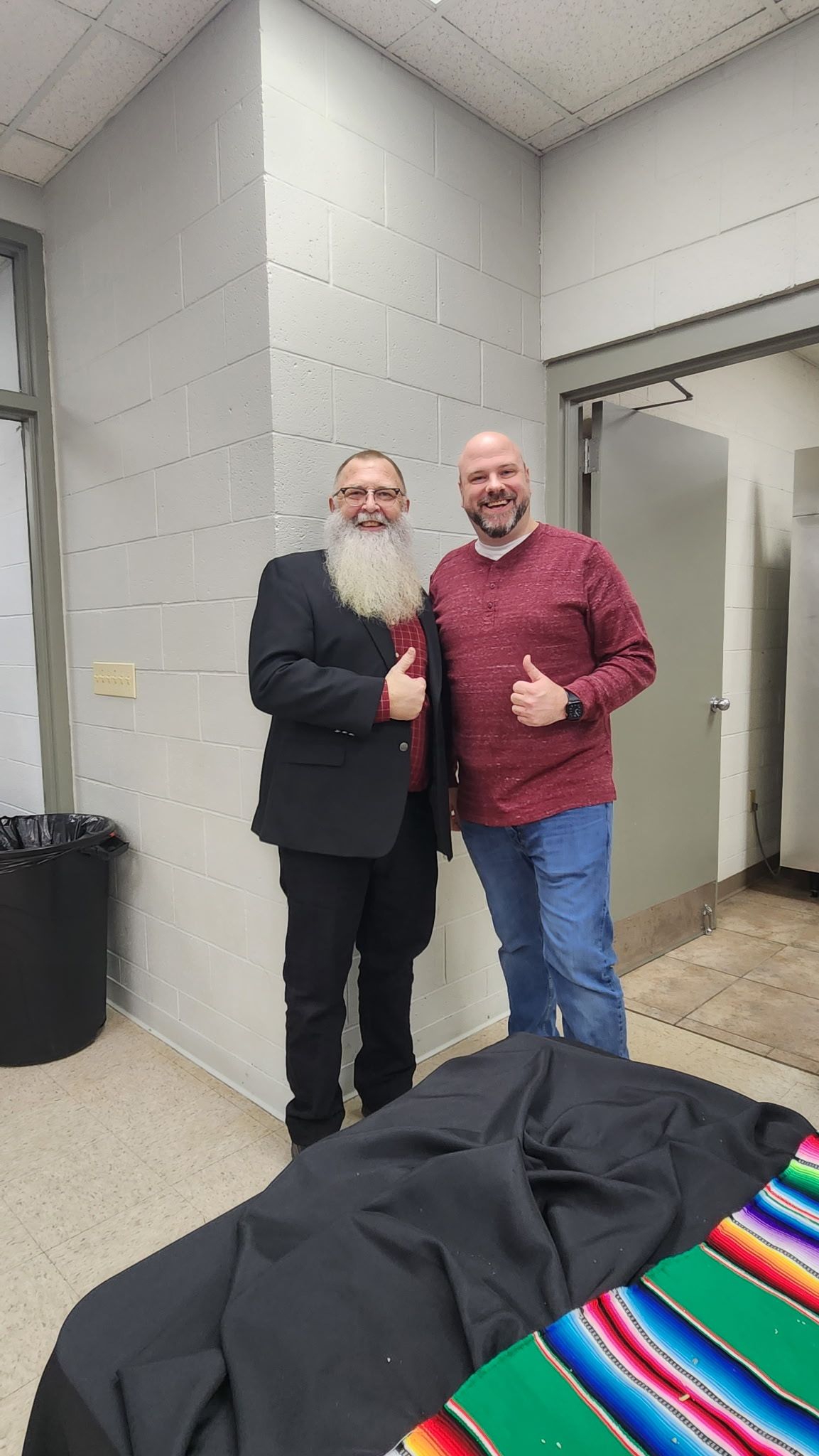 Two men giving thumbs-up. One with a long white beard, wearing a suit; the other in a red shirt and jeans. Inside a building.