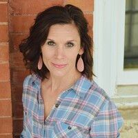 Woman with brown hair and earrings in blue plaid shirt smiling, near a brick wall and white door frame.