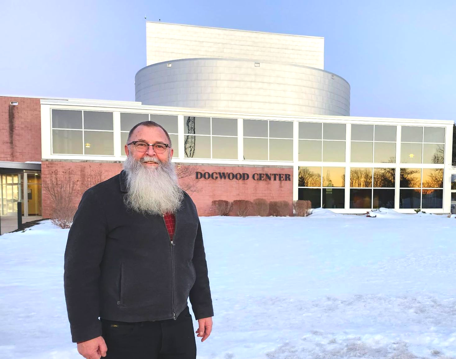 Man with long beard smiles in front of the Dogwood Center building, snowy ground, and blue sky.