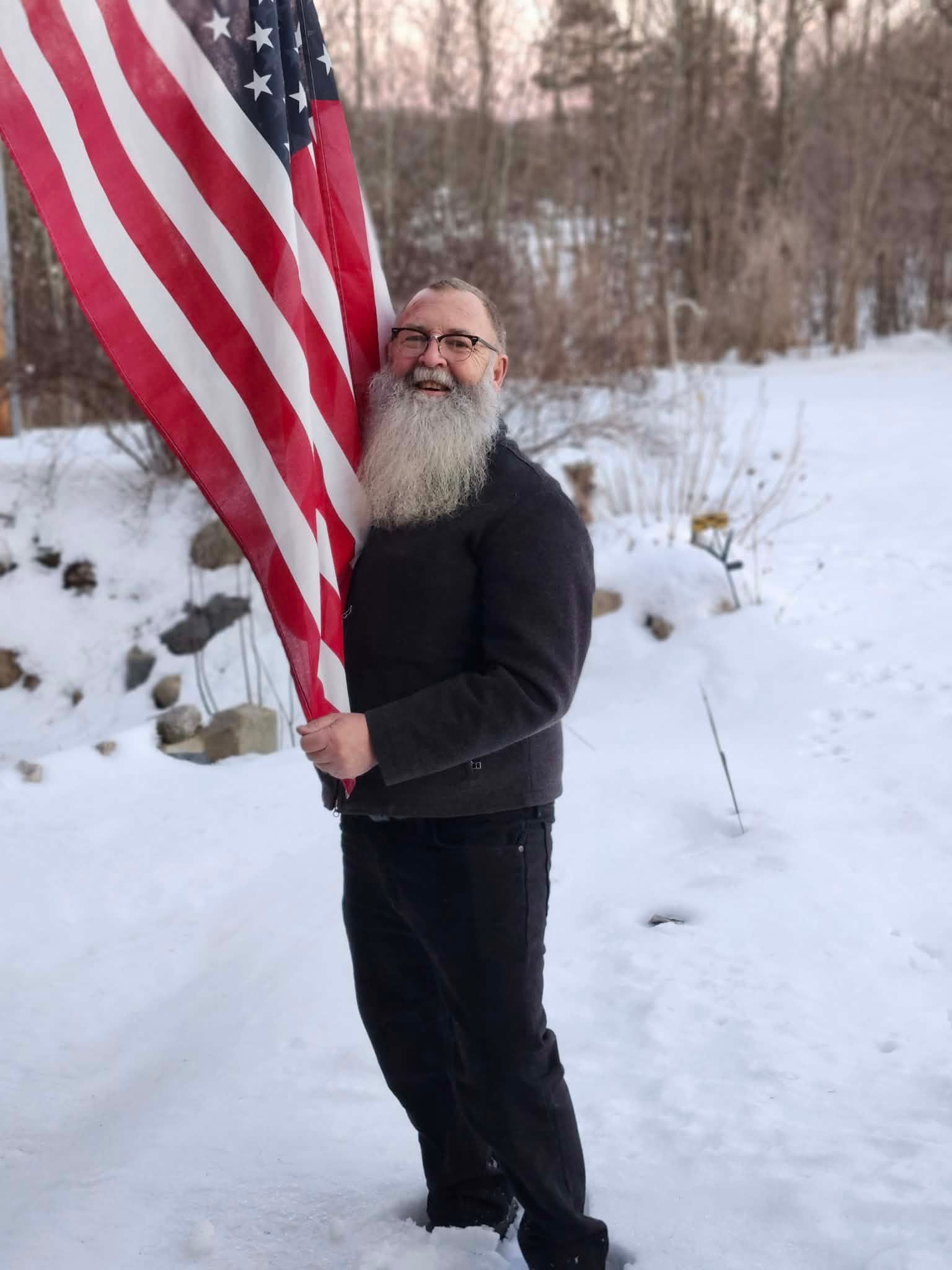 Man with long white beard holds American flag in snow.