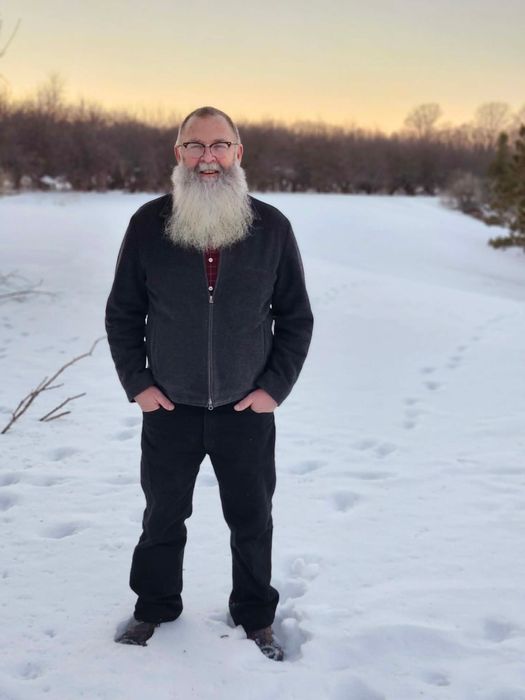 Man with long white beard stands in snow, hands in pockets. Trees in background.