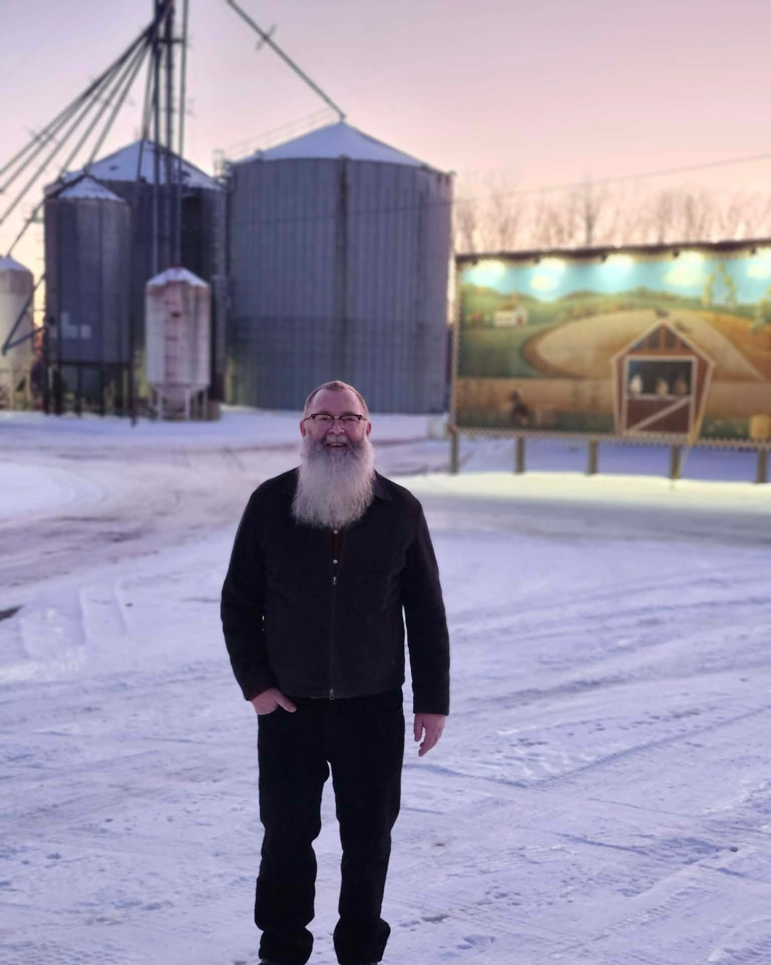 Man with long beard smiles in snowy field, silo and mural backdrop.