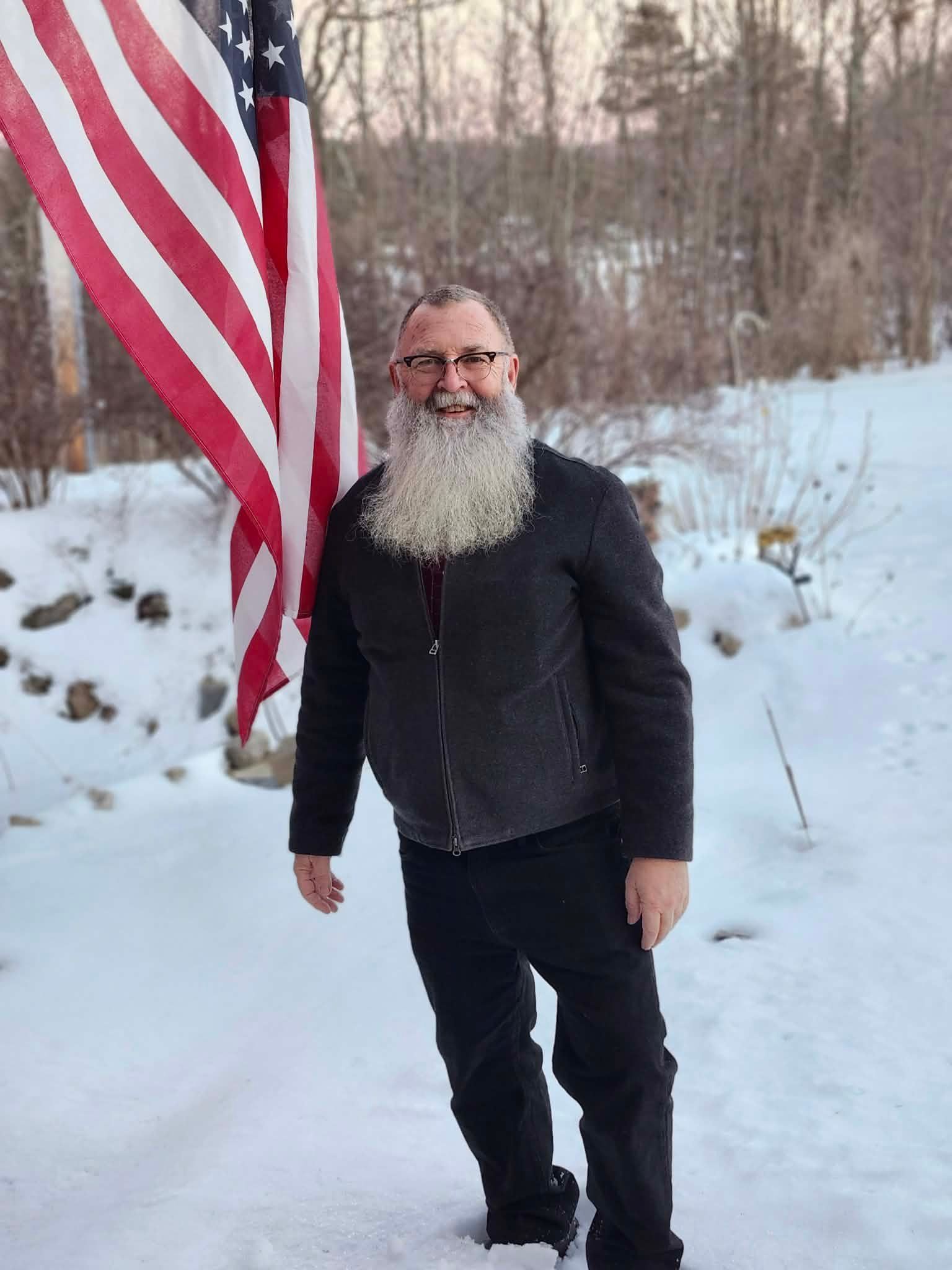 Man with large white beard stands in snow next to an American flag.