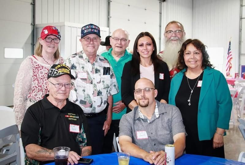 Group of people smiling, posing for a photo indoors, some wearing nametags, likely at an event with an American flag visible.