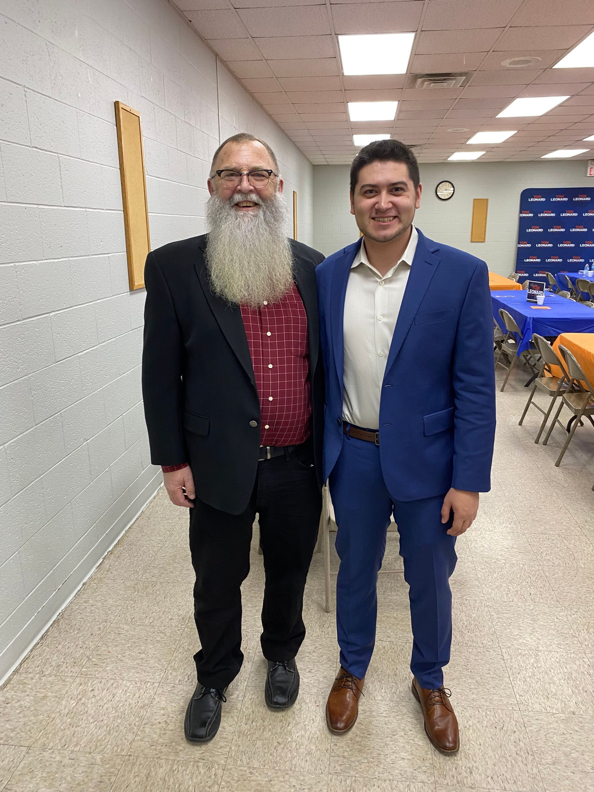 Two men pose indoors. One with a long beard in a black suit. The other in a blue suit. They smile.
