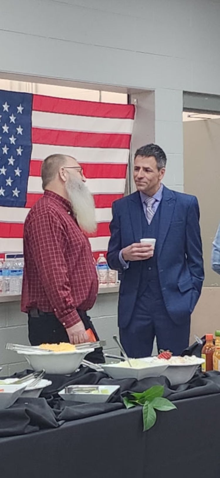 Two men converse near a buffet table with an American flag backdrop.
