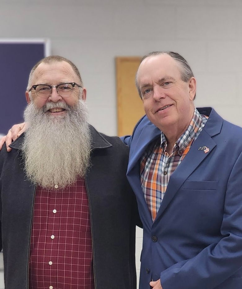 Two men smiling, arm around each other. Man on left has long beard, red shirt. Man on right in blue blazer, plaid shirt.