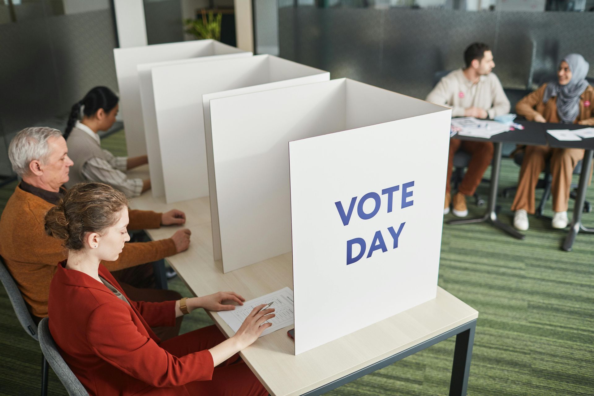 People voting at booths with volunteers at tables in a brightly lit room.